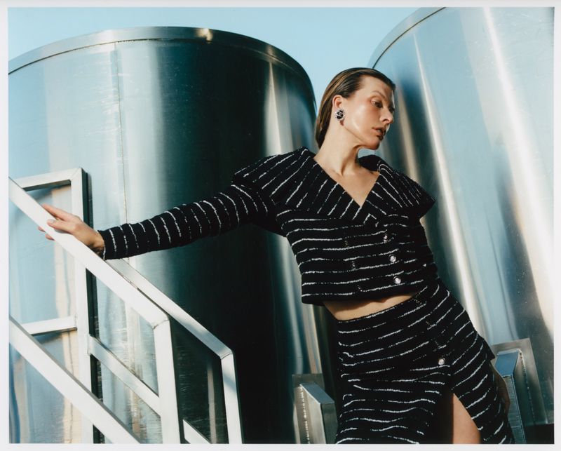 Fashion shot of an adult female posing outdoors in a striped jacket and skirt, standing near metallic tanks under a clear, light blue sky.