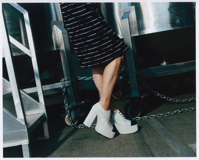 Model displaying a black and white striped skirt with white platform shoes. The setting is a factory or industrial environment.