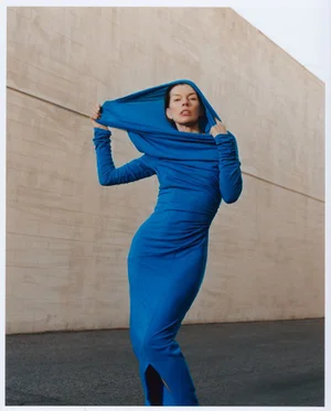 Milla Jovovich wearing a vibrant blue hooded dress standing against a concrete wall for Crash Magazine editorial photography.