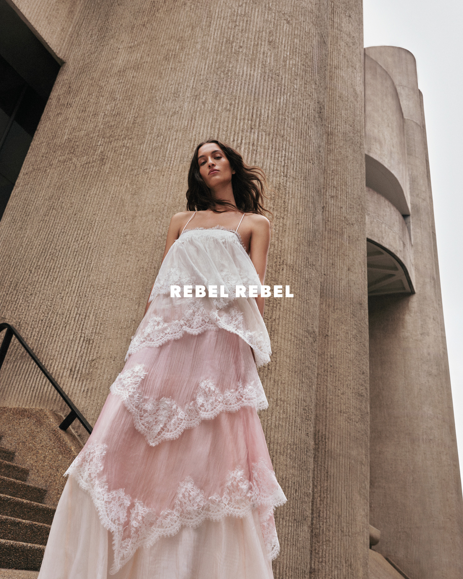 A woman poses in a layered pink-and-white gown against a concrete backdrop on a cloudy day, looking downward. The text 'REBEL REBEL' appears.