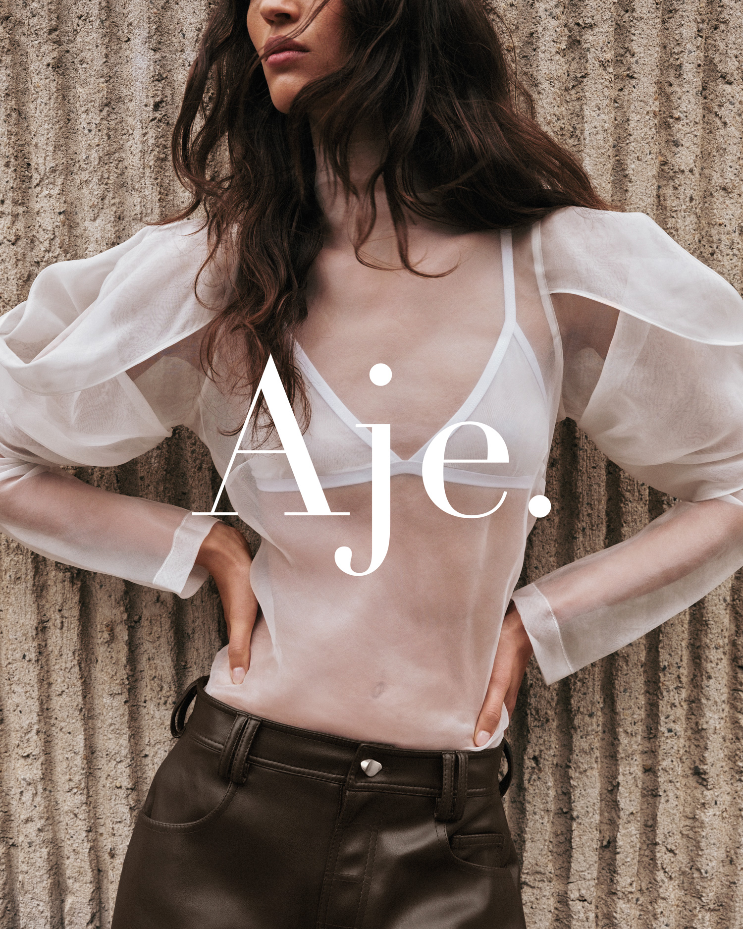 A woman poses in a studio with a sheer white blouse, brown leather pants, and a light concrete background for a fashion editorial.