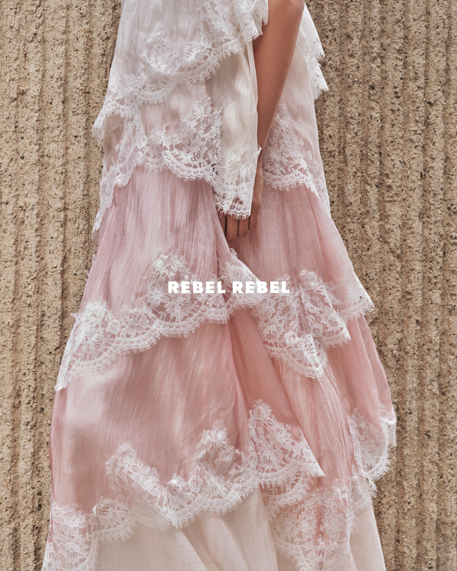 Close-up of a flowing, tiered gown in shades of pink and white with intricate lace detailing, textured background. Fashion editorial.