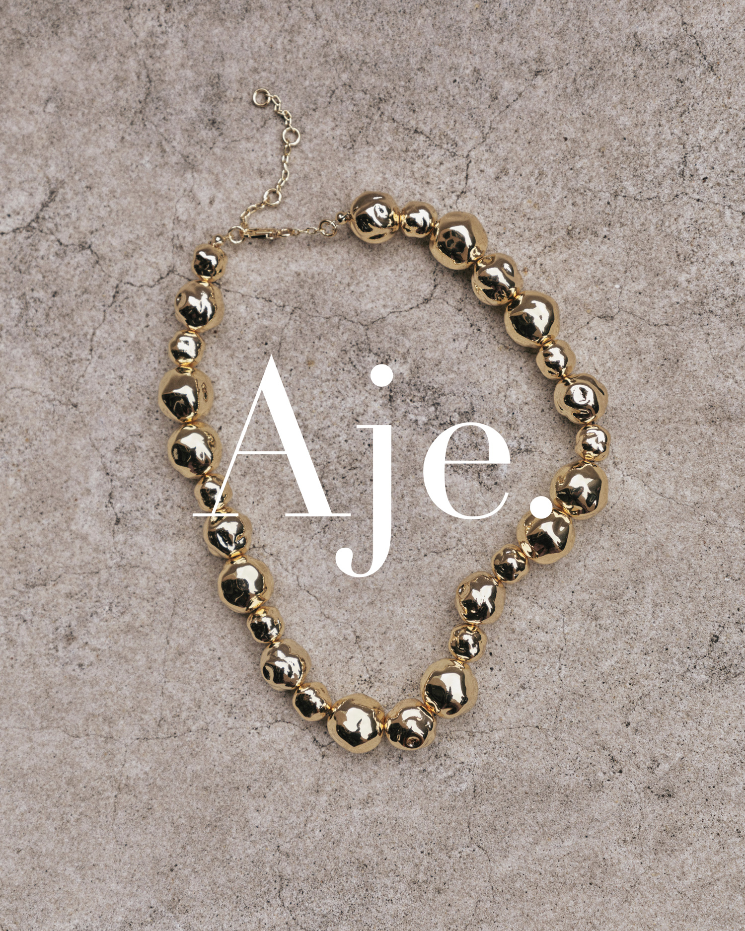 Gold beaded necklace on a textured grey surface with 'Aje' text overlay; product shot for fashion or jewelry design promotion.