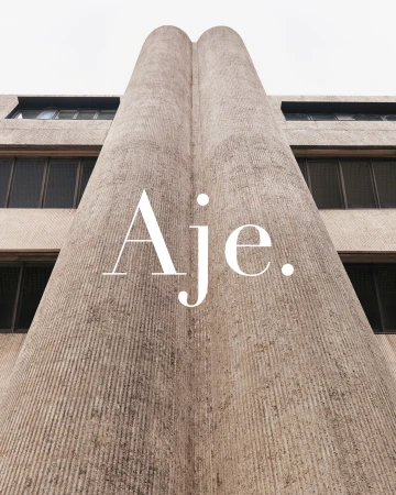 Low-angle shot of a textured concrete building facade with prominent vertical lines, windows, and the word 'Aje.' superimposed.