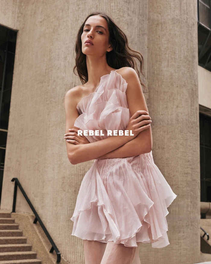 Model wearing a pink ruffled mini dress standing outdoors against a large concrete column and building exterior