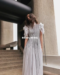 Model standing outdoors wearing a long white dress with black vertical embellishments near brutalist concrete architecture.