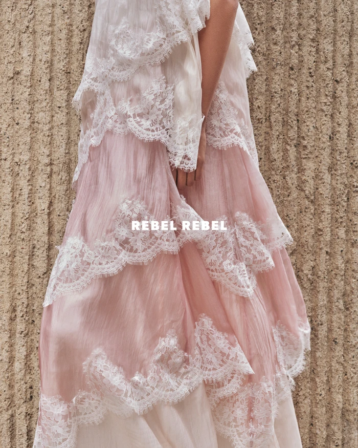 Model wearing a tiered pink dress with white lace overlay against a textured stone wall in a fashion editorial by Aje.
