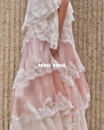 Model wearing a tiered pink dress with white lace overlay against a textured stone wall in a fashion editorial by Aje.