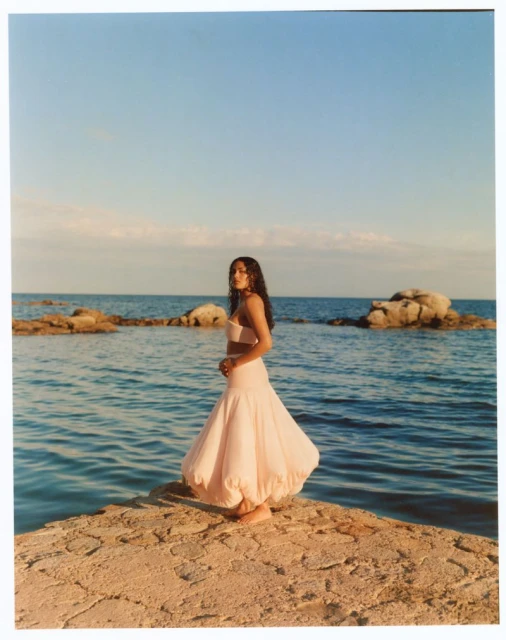 Adult woman in pink dress stands on a stone formation by the sea at daytime with a blue sky.