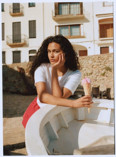 Woman with dark curly hair poses with ice cream cone, leaning against a white boat, with a building in the background outdoors.