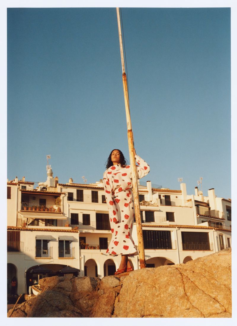 A woman in a patterned dress poses on rocks with whitewashed buildings and a blue sky in the background on a sunny day.