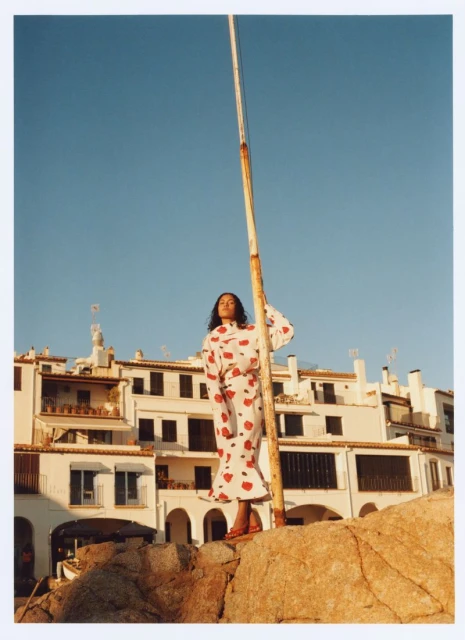 A woman in a patterned dress poses on rocks with whitewashed buildings and a blue sky in the background on a sunny day.
