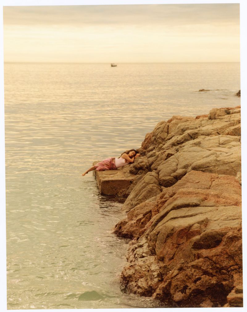 Woman relaxes on a rocky shore by the ocean, enjoying the sunset. A boat is visible in the distance against the horizon.