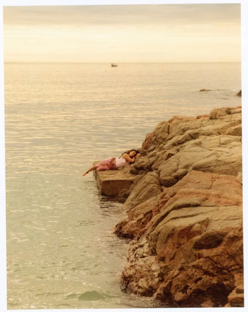 Woman relaxes on a rocky shore by the ocean, enjoying the sunset. A boat is visible in the distance against the horizon.