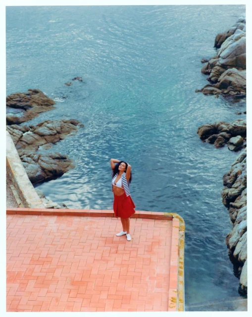 Adult female poses on a red tiled patio overlooking the turquoise sea, rocks, and sky, wearing a skirt, sandals, and striped top.