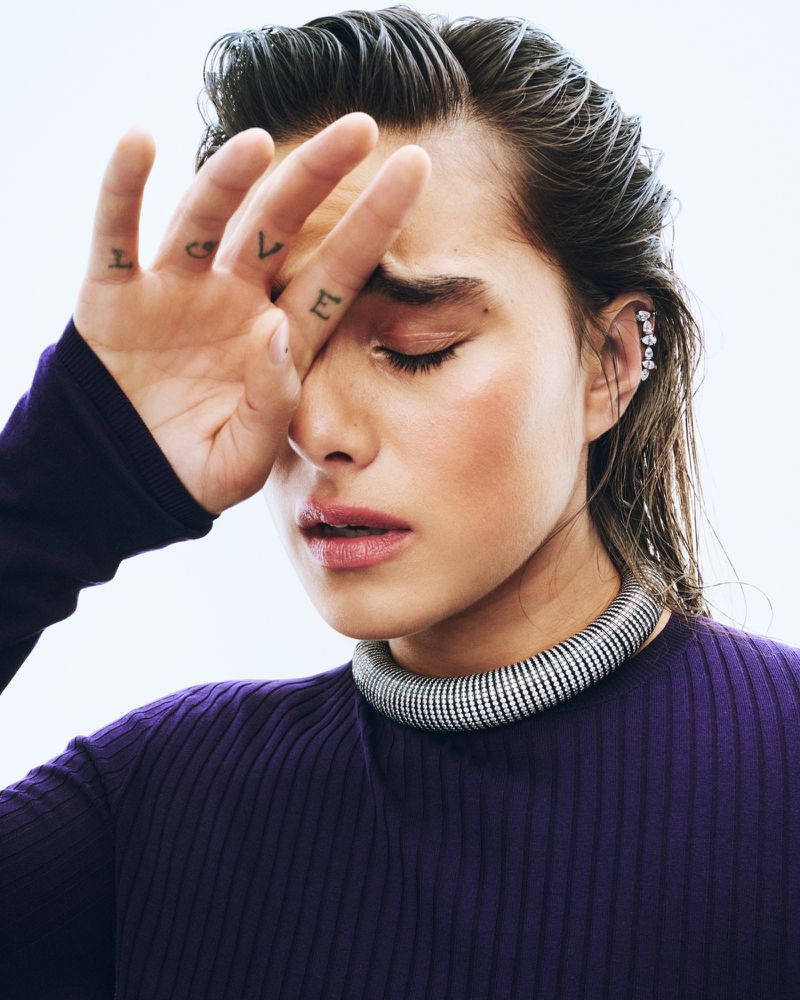 Close-up of a person with the word 'LOVE' tattooed on their fingers, posing with their hand near their face in a studio setting.