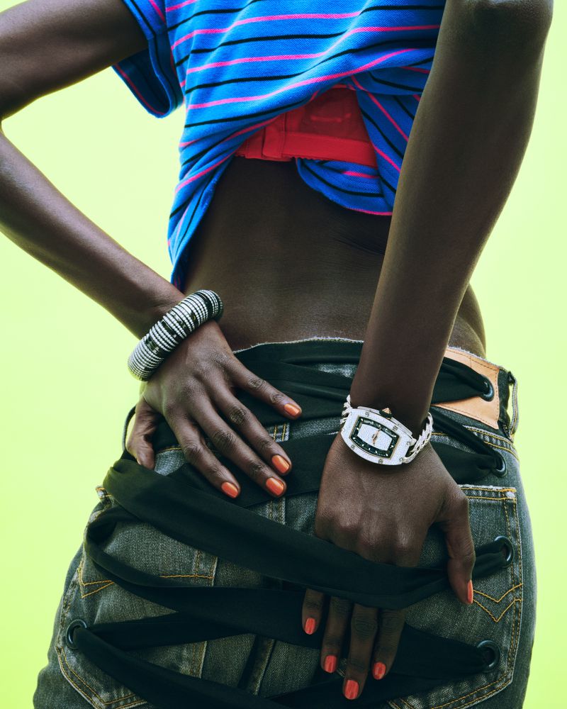 Dark-skinned woman in a striped shirt, jeans with ribbon detail, and jewelry poses against a light green background in a studio shot.