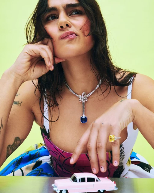 Woman poses with a necklace, ring, and toy car against a vibrant yellow backdrop. The jewelry and car are prominently featured.