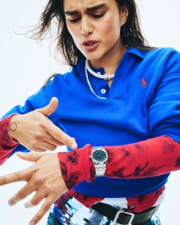 Portrait of a woman wearing a blue polo shirt, a silver watch, layered tops, and jewellery, shot in a photo studio against a white background.