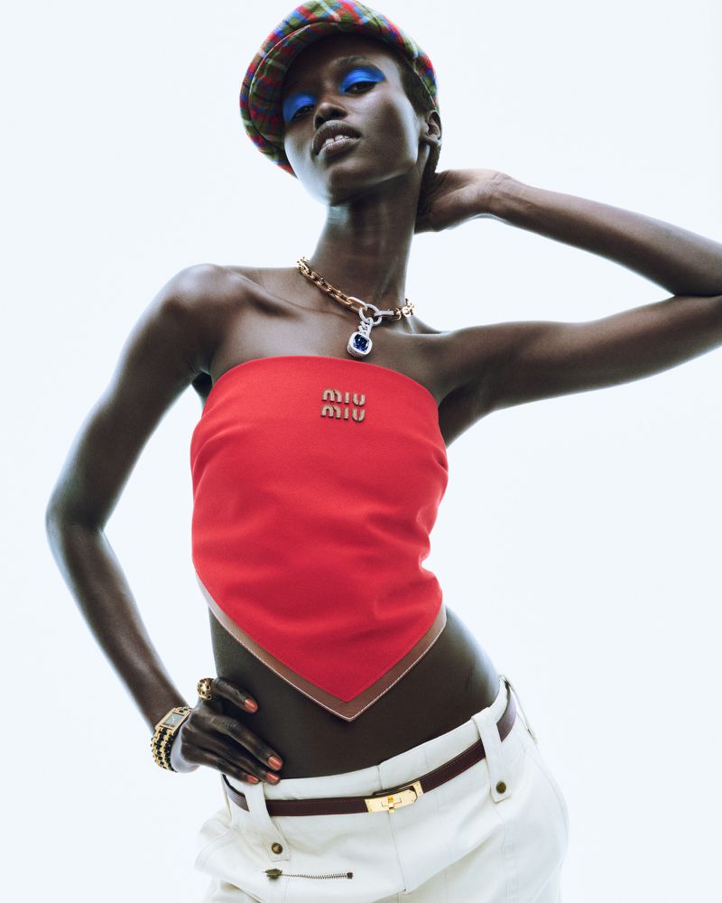 Fashion shot of a dark-skinned adult female wearing a red bandeau top with a logo, white pants with a brown belt, and a plaid beret.