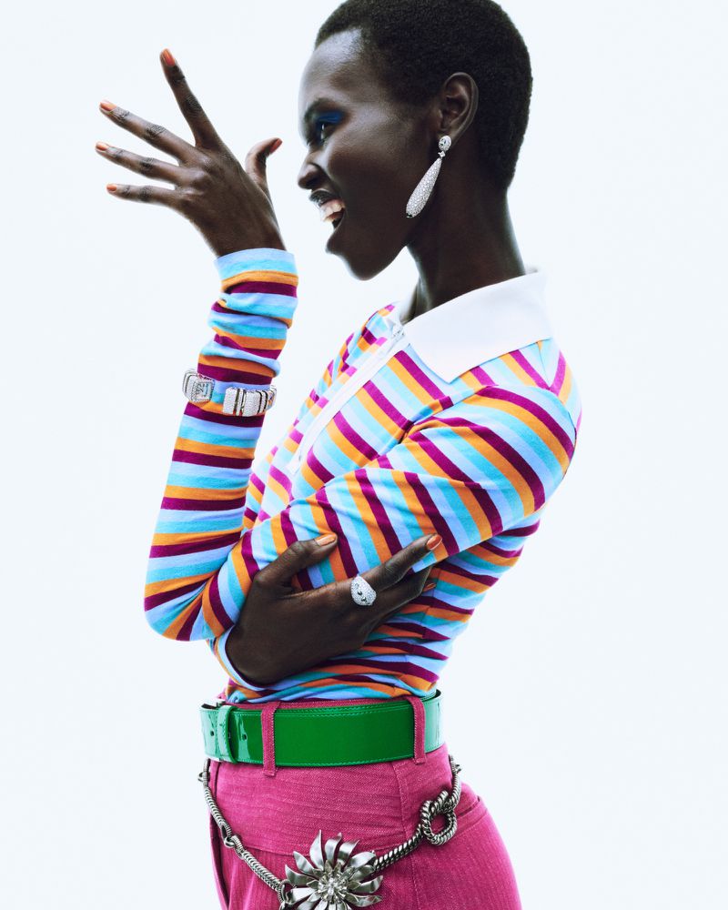 A stylish young Black woman poses in a colorful striped top, pink pants, and statement jewelry against a bright white backdrop.