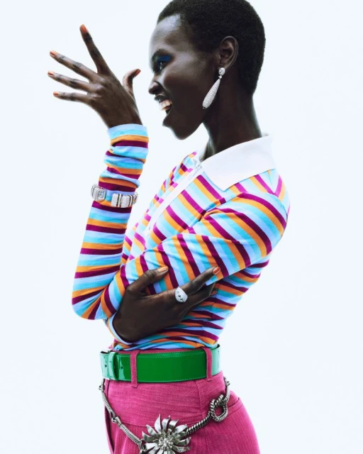 A stylish young Black woman poses in a colorful striped top, pink pants, and statement jewelry against a bright white backdrop.