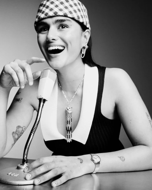 Smiling woman in a checkered hat sits at a table with a vintage microphone, wearing jewelry. Black and white studio portrait.