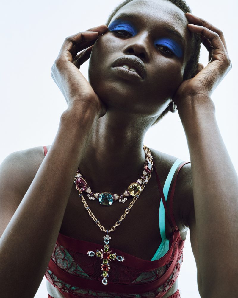 Close-up of a woman with dark skin and blue eyeshadow, wearing an elaborate beaded necklace with colorful jewels, posing indoors.
