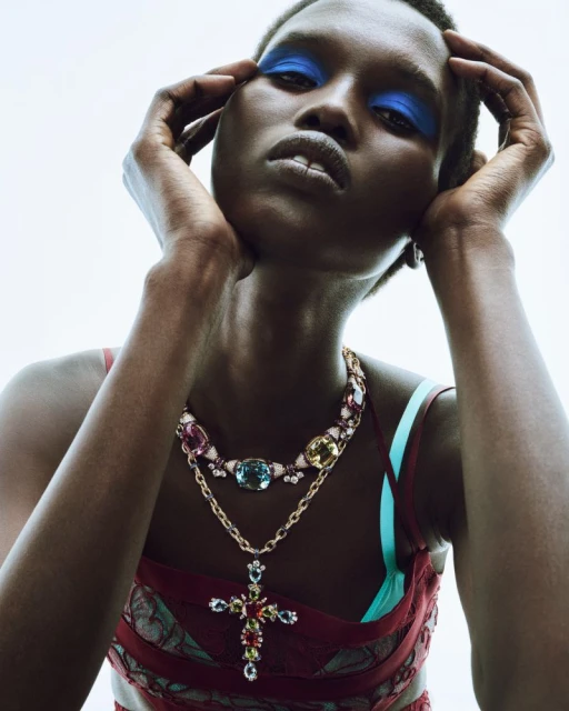 Close-up of a woman with dark skin and blue eyeshadow, wearing an elaborate beaded necklace with colorful jewels, posing indoors.