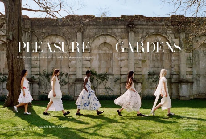 A fashion editorial shot outdoors featuring five women in long white dresses walking on green grass in front of an old stone wall.
