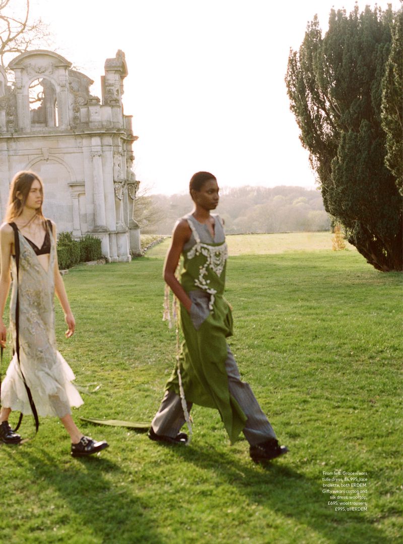 Two fashion models walk across a grassy field with an old stone building and trees under sunlight. They wear loose dresses.