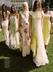 Five female models in flowing pastel dresses standing outdoors on a green lawn in front of a stone building.