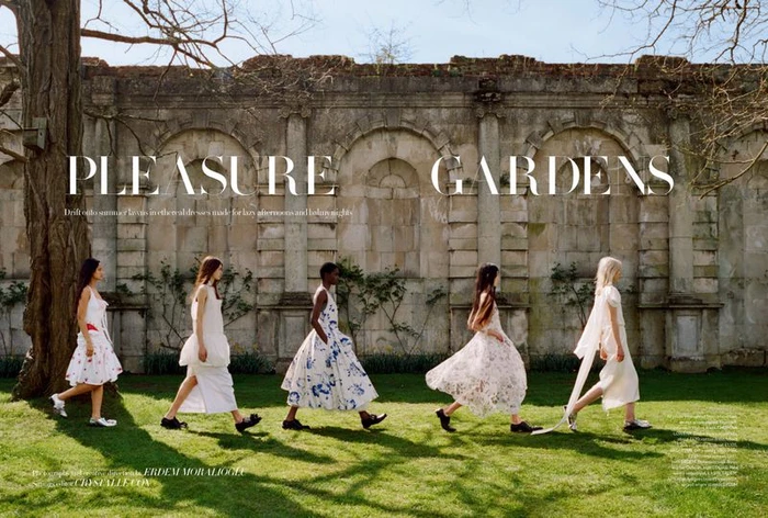 Five models in flowing white dresses walking across a grassy lawn before an ancient stone wall