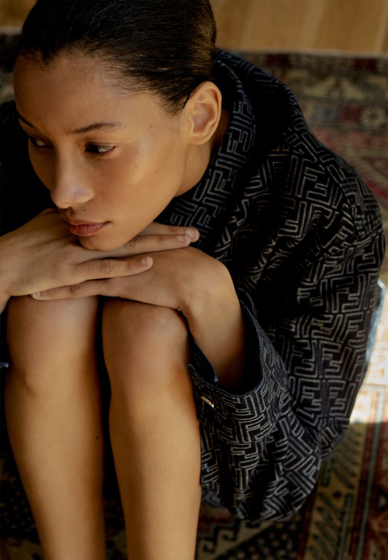 Young woman in patterned jacket sits with knees bent and hands clasped; indoor setting, warm lighting, portrait-style frame.