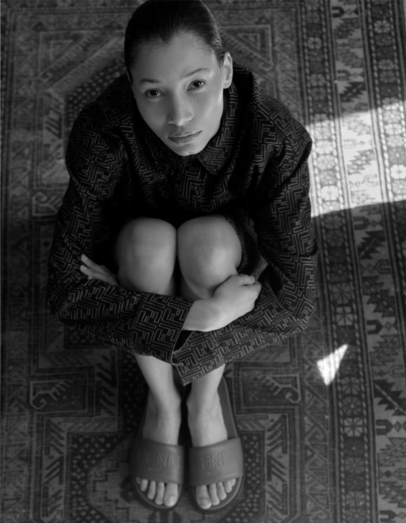 Overhead portrait of a woman kneeling on a patterned rug wearing a patterned shirt and sandals in a brightly lit indoor space.