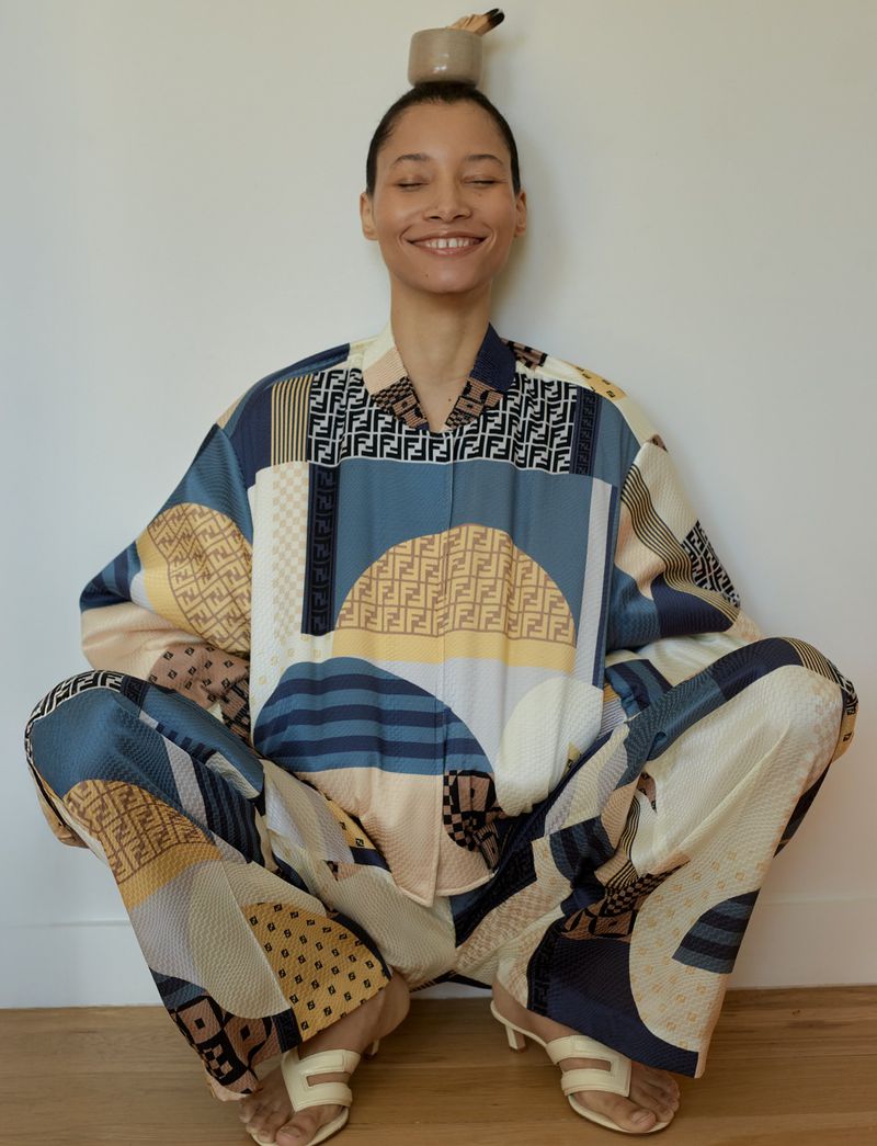 A woman in patterned pajamas balances a small pot on her head, smiling against a white wall, indoors. The shot is eye-level.