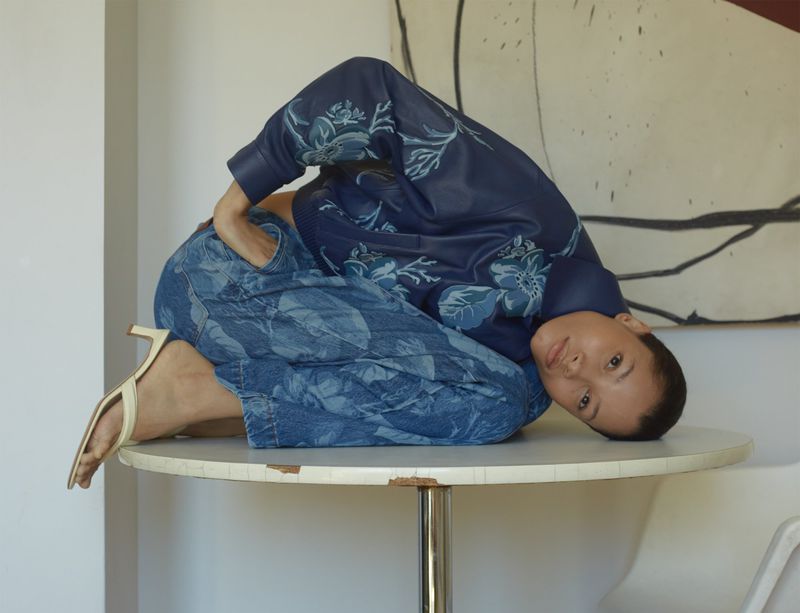 Woman in blue floral jacket and pants poses creatively on a white round table with light background and soft lighting in a studio.