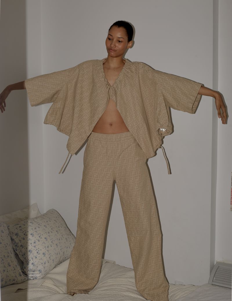 A woman in a beige linen two-piece outfit stands on a bed in front of a white wall, arms outstretched, captured in a medium shot with soft lighting.