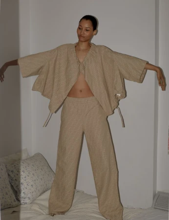 A woman in a beige linen two-piece outfit stands on a bed in front of a white wall, arms outstretched, captured in a medium shot with soft lighting.