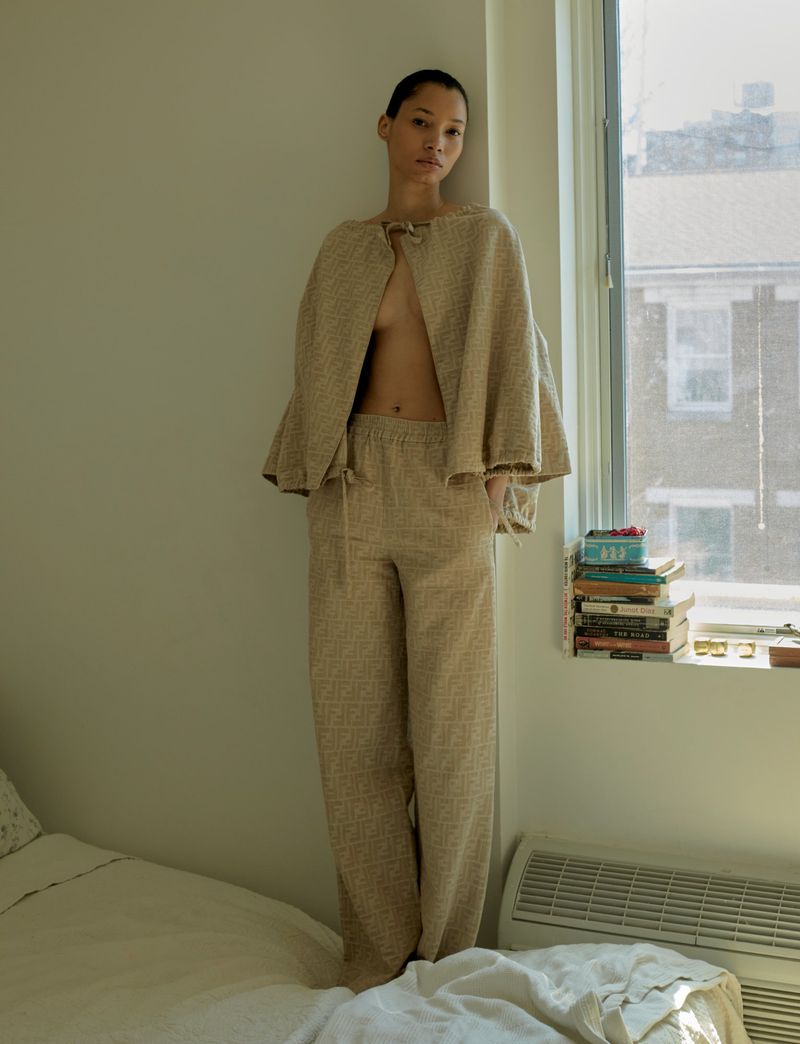 Woman in neutral-tone lounge wear posing in an indoor room with natural light from a window, books, and a radiator behind her.