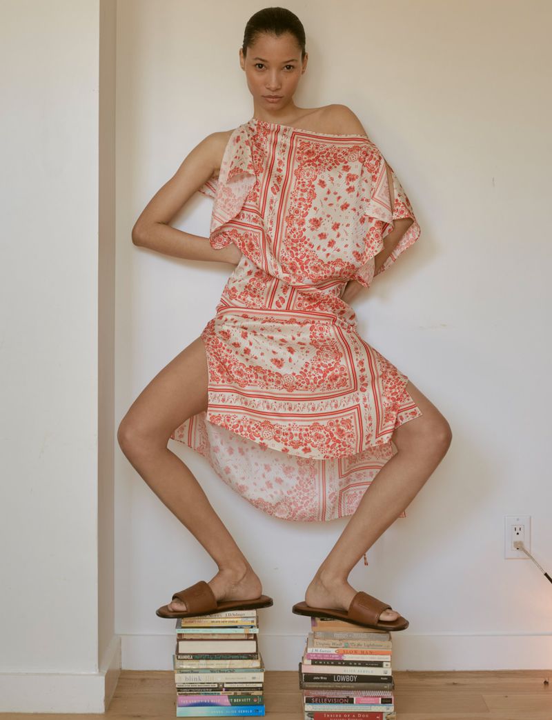 An adult female poses indoors, wearing a patterned dress and sandals, standing atop stacked books in front of a plain wall.