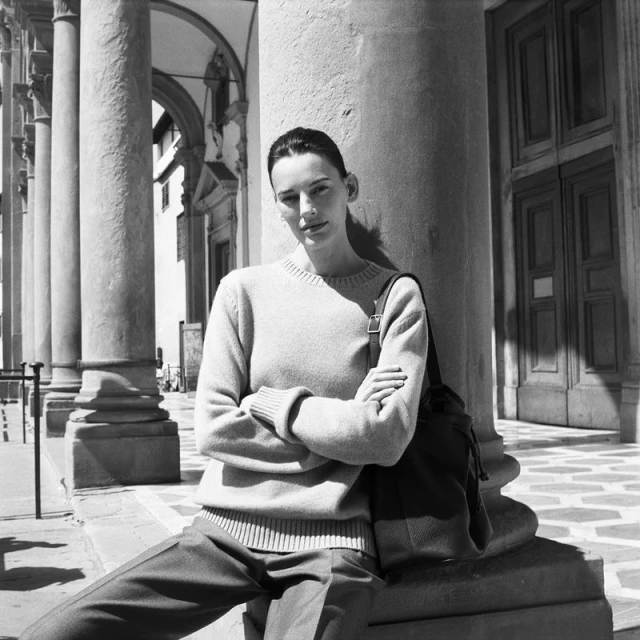A woman in a sweater and pants poses outdoors near columns. She has her arms crossed and is standing next to a column.