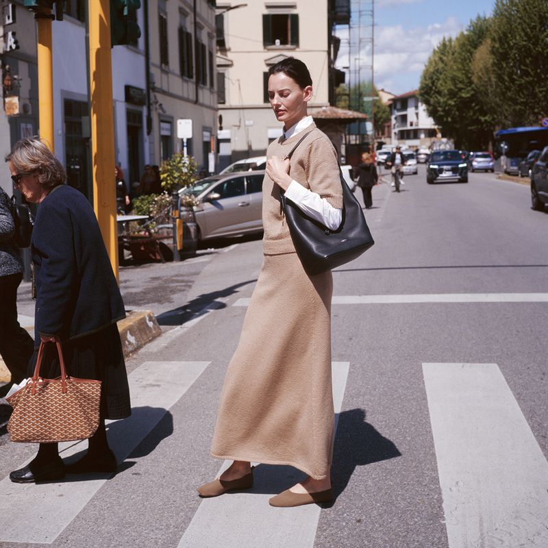 Woman in tan skirt and sweater walking across a crosswalk in an urban setting, with cars and buildings visible in the background.