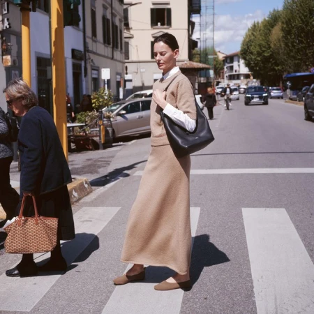 Woman in tan skirt and sweater walking across a crosswalk in an urban setting, with cars and buildings visible in the background.