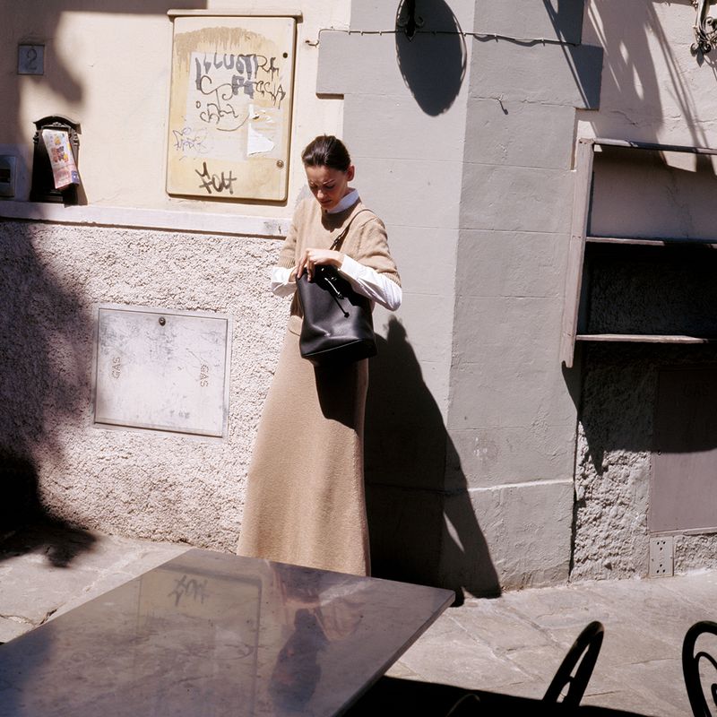 Adult woman in a beige dress and white shirt opens a black purse next to a table on a city street on a sunny day.