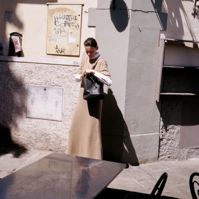 Adult woman in a beige dress and white shirt opens a black purse next to a table on a city street on a sunny day.