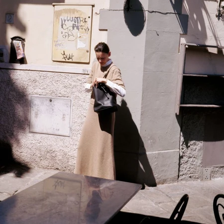 Adult woman in a beige dress and white shirt opens a black purse next to a table on a city street on a sunny day.