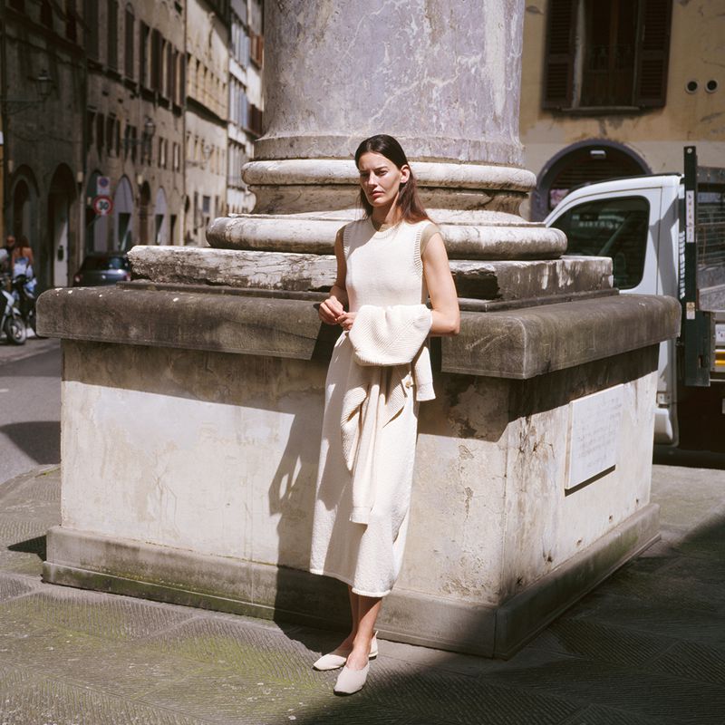 A brunette woman in a light cream dress stands near a marble column in a city, holding a matching sweater, with soft, natural lighting.