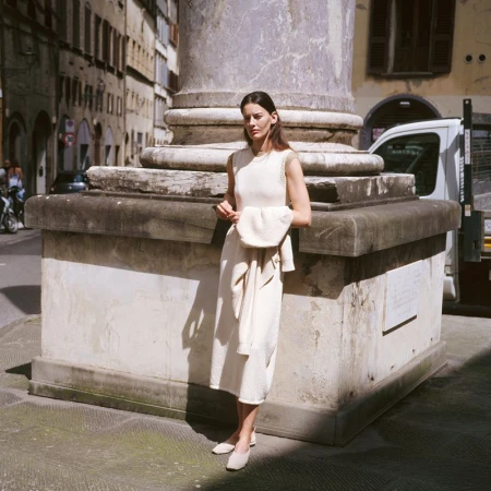 A brunette woman in a light cream dress stands near a marble column in a city, holding a matching sweater, with soft, natural lighting.