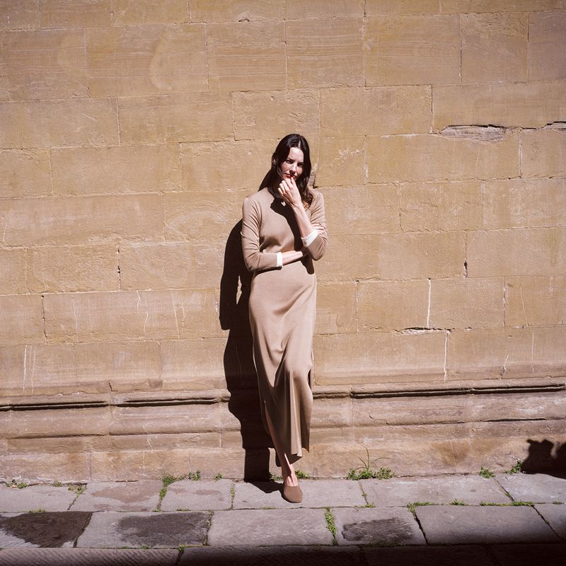 A woman in a tan dress stands against a textured wall. She is in full shot with her hand near her face.